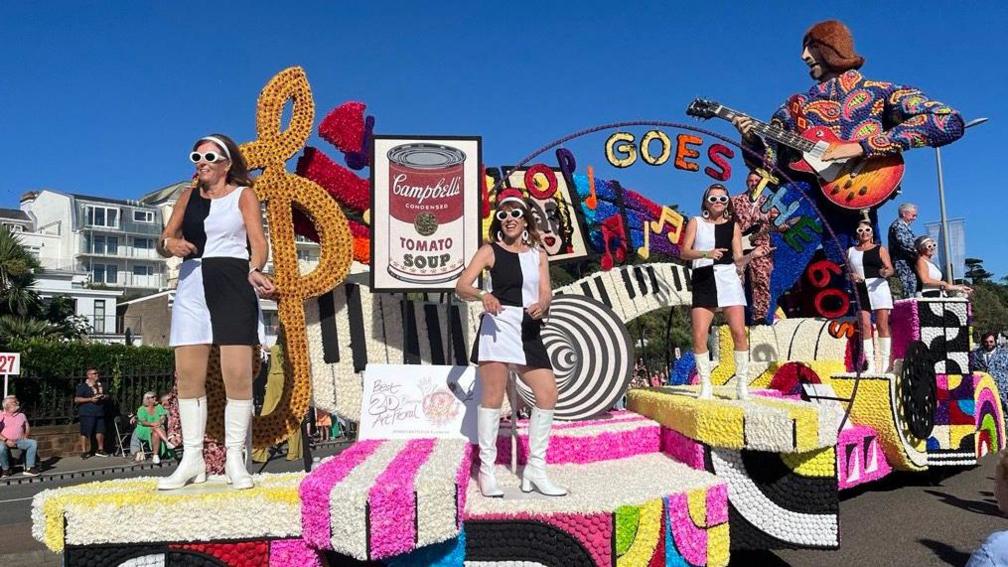 a float themed around the 1960's it include a large sculpture of a man in a floral shirt playing the guitar and bright psychedelic colours 