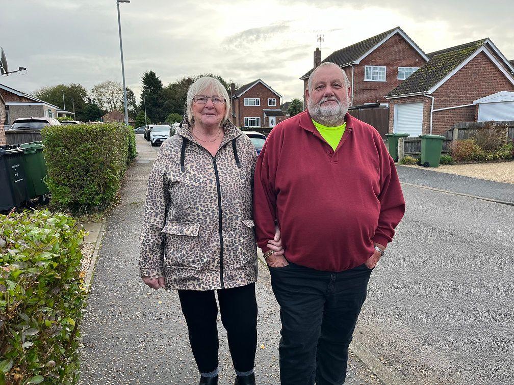 Sue and Tony Bojko stand outside Toftwood Medical Centre. Sue is wearing a leopard print coat and black leggings. Tony is in a red top with a bright yellow T-shirt under it and dark blue trousers. He has his hands in his pockets, but she has wrapped her hand under his arm and is holding his wrist. 