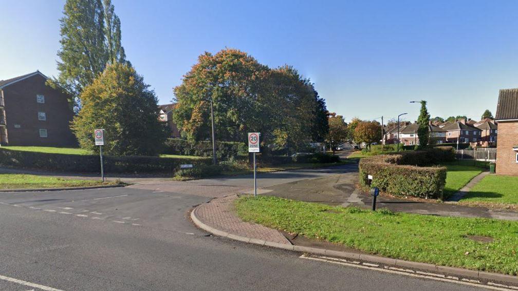 A road in Rotherham showing the entrance to a housing estate and two 20mph signs in view, as well as several trees and gardens 