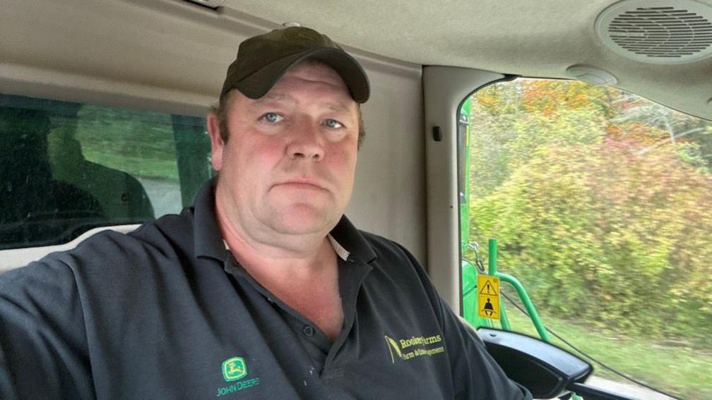 Dan Willis is pictured at the wheel of a tractor or other large vehicle. He wears a dark baseball cap and a grey top.