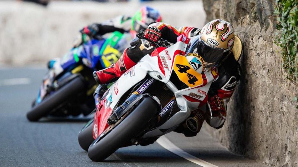 Riders leaning into a stone wall on a bend, the bikes and leathers are colourful and the bike at the front at the number 44 in black on a yellow plate on the front.