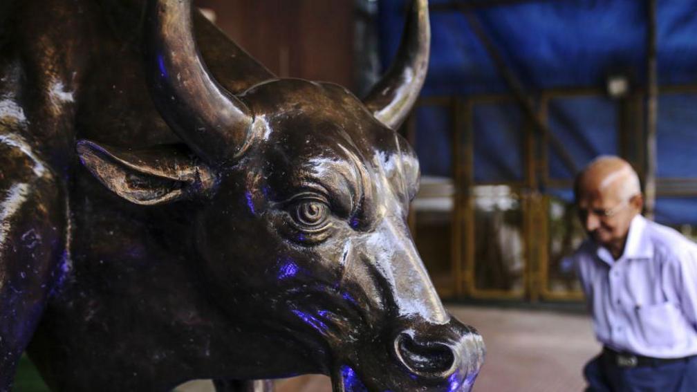 A bronze bull statue stands at the entrance to the Bombay Stock Exchange, with a balding man walking past it.