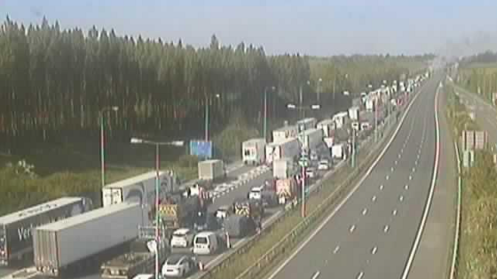 National Highways CCTV footage showing traffic building up toward the fire, with cars and lorries stuck in standstill while emergency services work on ensuring people's safety. The sky is clear and blue with smoke rising up on the horizon in the background.