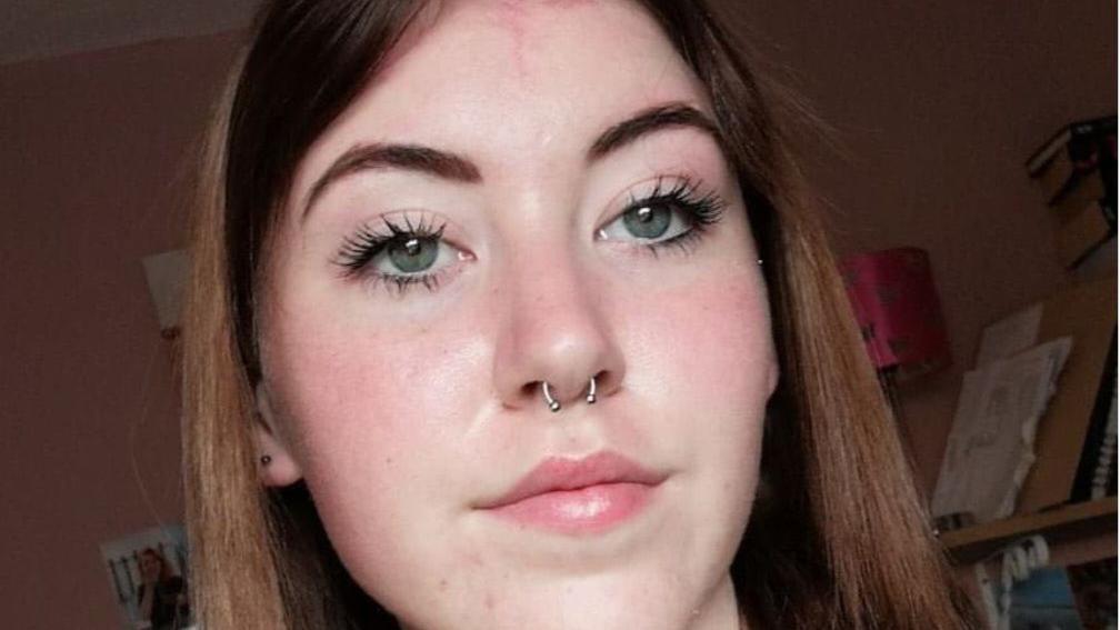 A teenage girl with should length, brown hair. She has a pale complexion and is wearing a nose ring.