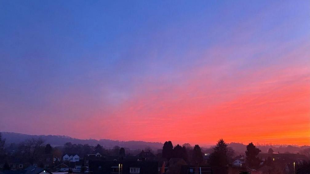 A half blue sky is shot through with a big chunk of red and orange colours, over the roofs of houses and trees