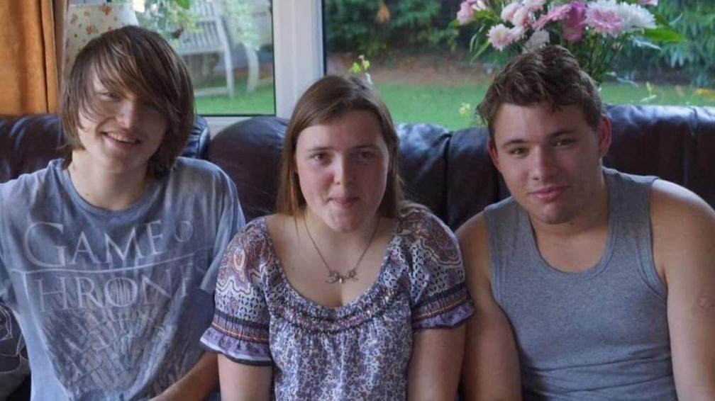 Two boys sit either side of Holly on a black leather sofa. Stephen is sat on the left with a faded blue Game of Thrones t-shirt, Holly is sat in the middle with short brown hair, a necklace and floral blue dress, with Dale on the right wearing a grey vest.