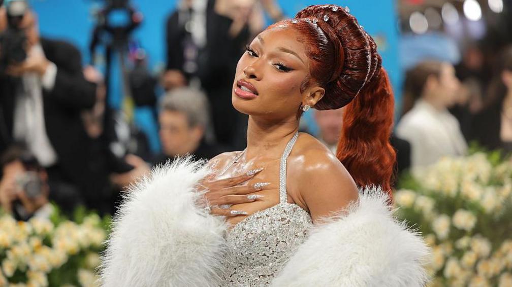 A woman wears a strappy, sparkly A-line gown. Her hair is styled in an elaborate updo with a long, red pony tail spilling down her back. She sports long, pointy silver nails decorated with a sparkly design, and a white, fluffy stole draped over her shoulders.