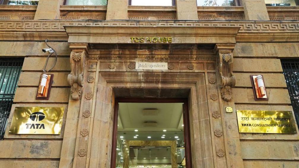 The image is of the Mumbai headquarters of TCS. It is an ornate colonial building in brown stone, with golden plates of TATA and Tata Consultancy Services displayed on either side of the main door.