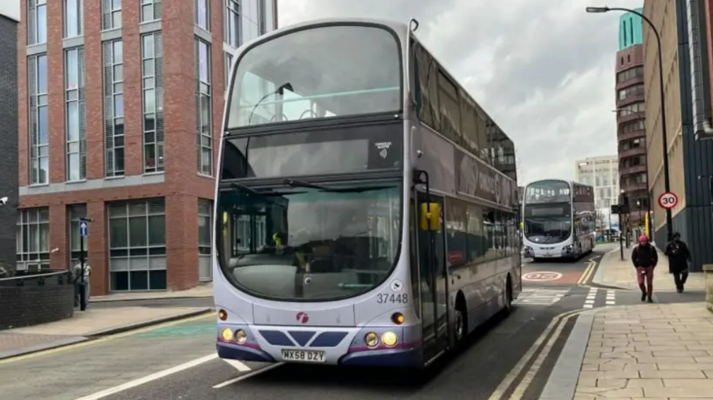 A pale blue double-decker bus being driven along a city centre street.