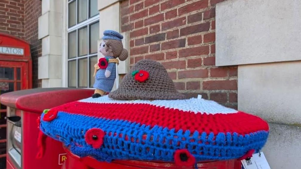 The image show a knitted Remembrance Day post box topper in blue, red and white with a helmet with a poppy on it and a female character in uniform. There is a space to the right hand side where the knitted soldier had been.