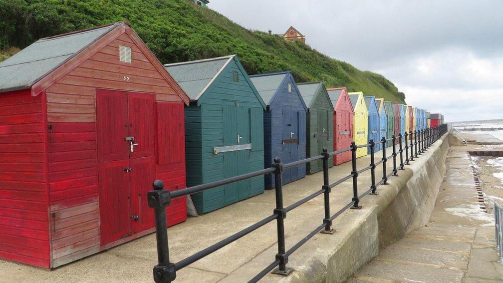 A row of brightly-coloured beach huts lined along a concrete promenade, with the sea wall and railings to the right and a grassy steep slope behind.