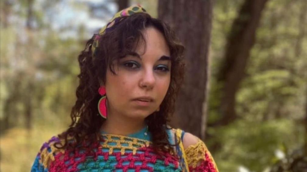 A woman wearing a multicoloured jumper and bright pink earrings with long brown hair stands in a forest.