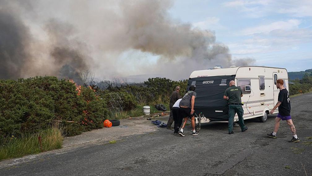 A caravan being moved as flames lick roadside vegetation