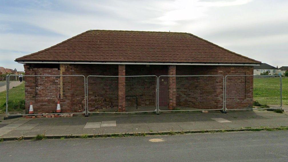 A one-storey brick-built building with a brown tiled roof, surrounded by metal fencing, a pathway and a grassy area