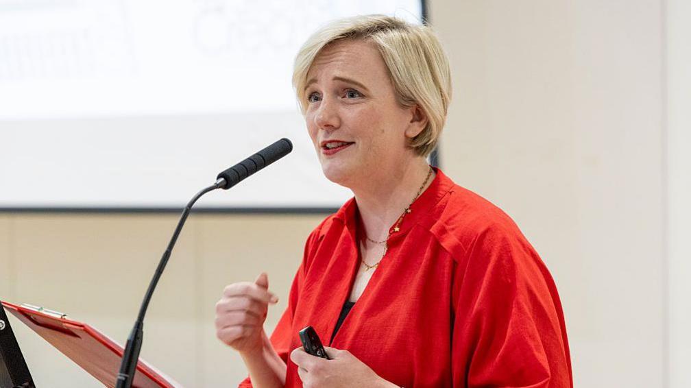 Creasy in a bright red top speaks at a lectern while holding a presentation remote. A microphone is positioned in front of her.