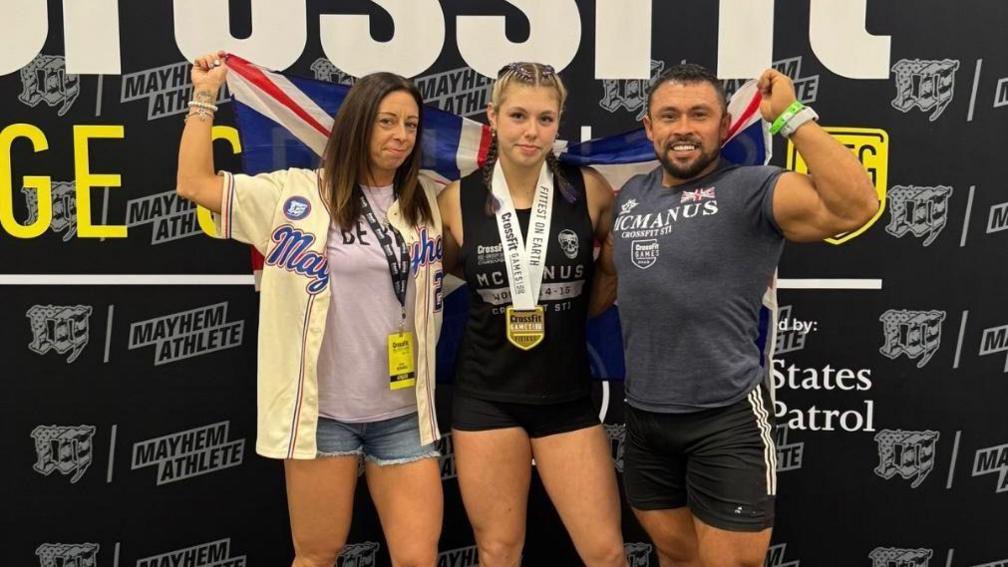 Keira, standing in between her mum Lisa and dad Joel who are holding a union jack flag, and has a medal around her neck. They are in front of a step and repeat board, with the word Crossfit and various sponsorship logos on it.