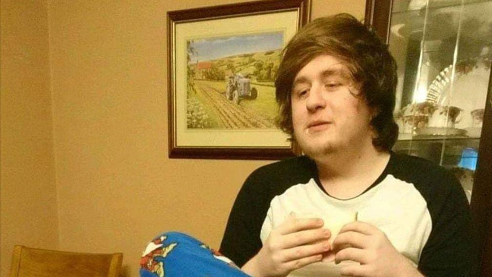 Alexander McCartney with long brown hair looking away from the camera, with a yellow wall and painting in the background