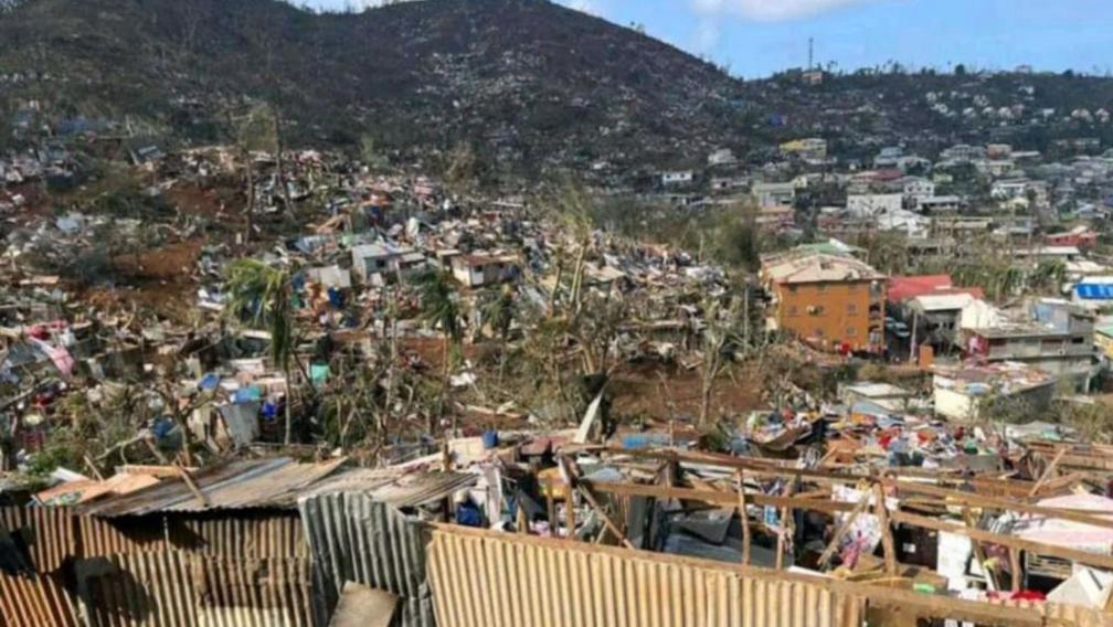 Damage in Mayotte. A town is seen in ruins, with a hill scape rising in the background. Trees appear blown over, walls are collapsed, though some buildings remain standing.