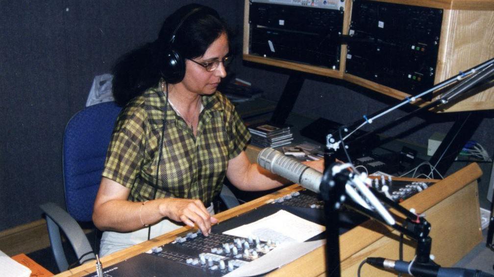 A woman wearing headphones sits in front of a microphone at a radio desk in the 1990s.