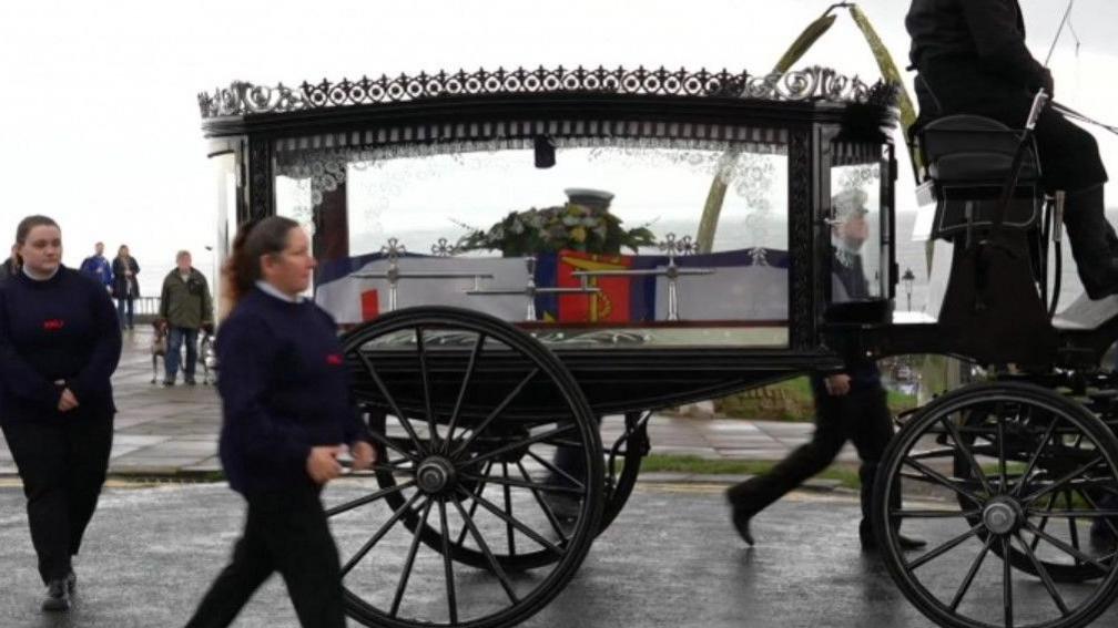 A horse-drawn glass hearse on a road. Inside the hearse, a coffin is draped in a Union Jack flag with a floral arrangement on top. Two women in dark uniforms walk alongside the hearse and the driver is seated at the front, dressed in black. In the background, several onlookers are standing. 