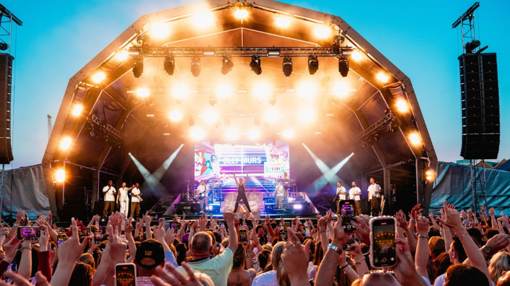 A picture taken form the crowd of Olly Murs on the stage dressed in a short sleeved black top and black trousers with his arms up in the air in a v shape, and an Olly Murs written backdrop. There are three brass instrumentalists on the right of the stage (his left), dressed in dark trousers and white shirt tops. There are three backing vocalists on the left (Olly's right), with the middle singer dressed all in white, and the two either side in dark trousers and white shirts. The canopy of the stage has multiple lights and you can see the sky darkening in the background.
In the foreground of the picture you can see maybe a couple of hundred people with arms in the air, many holding phones and seemingly recording Olly's set.