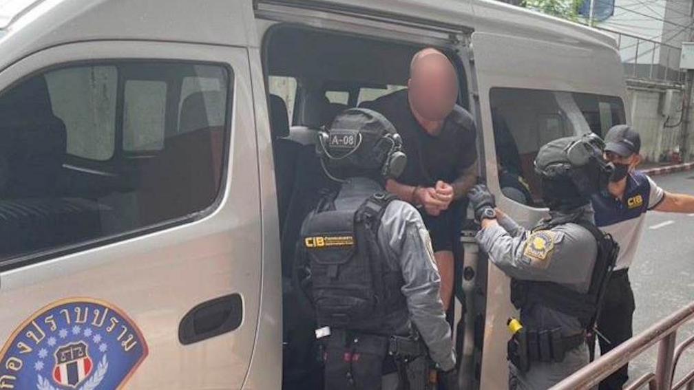 A man with his face blurred walking out of a Thailand Police car van with two Thai Police officers standing in front of him. The man is wearing a black top with his hands in front of him.