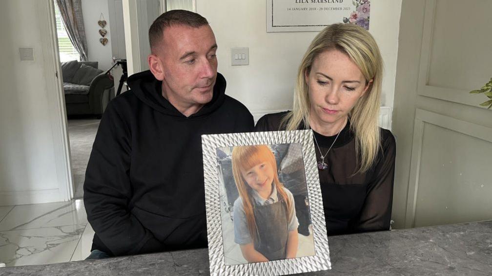 Lila's parents are looking at photograph of Lila in a silver frame. They are both sitting on stools in their kitchen.