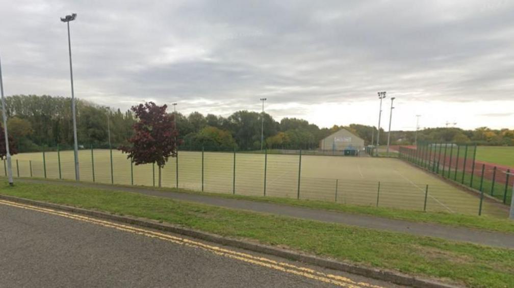 An image of an artificial football pitch surrounded by fencing.  It is bordered by trees to the left. There is a house in the background and an athletics track to the right. Floodlights on stands are visible in the background.