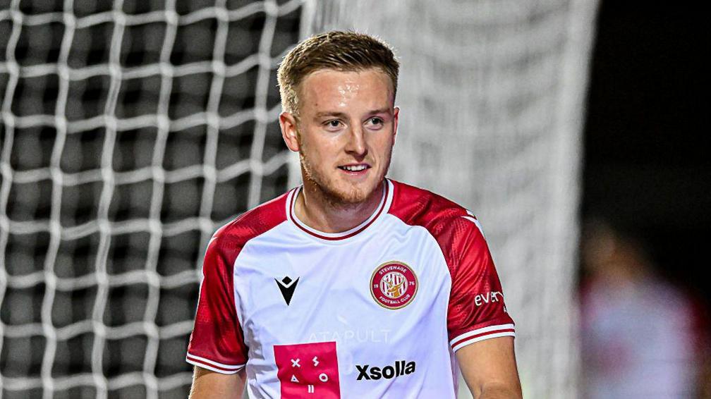 Harvey White smiles looking ahead with sweat on his brow, wearing home Stevenage kit of white and red