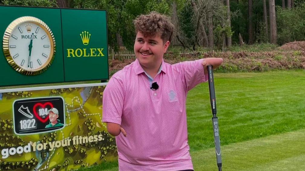 Adam has under-developed arms. He is holding onto a golf club with his arm and wearing a pink golf shirt.