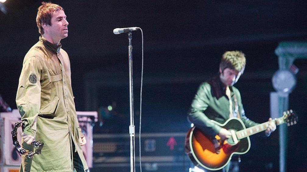 Liam and Noel Gallagher stood on stage. Liam is wearing a green Parka coat and holding a tambourine, Noel is playing a guitar.