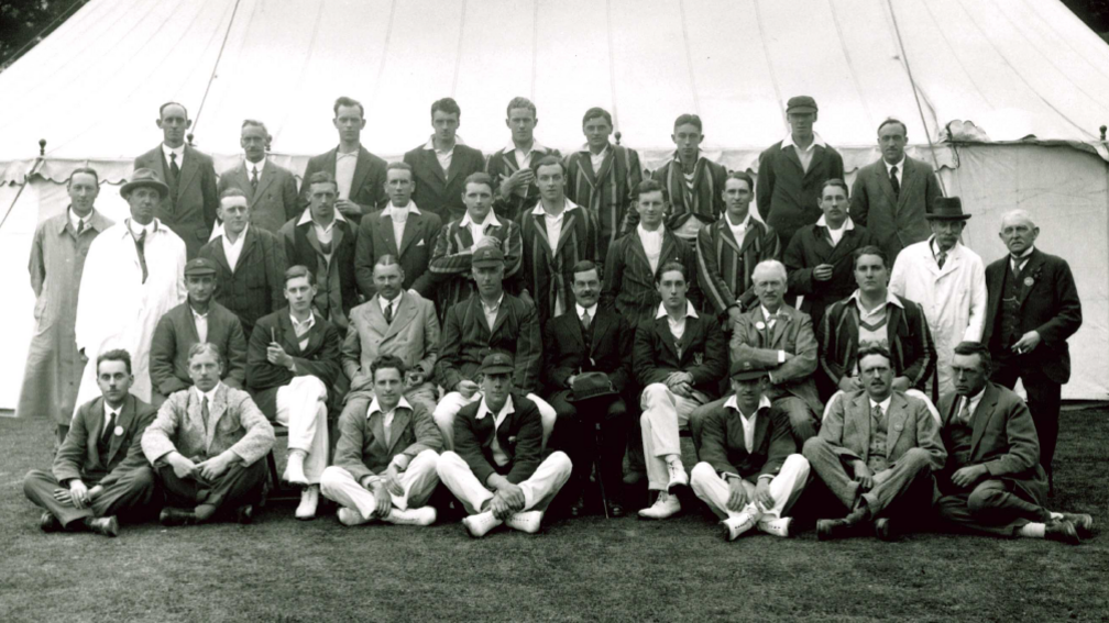 A black and white team photo taken in front of a large white tent. The male players are either sitting or standing across four rows. They are wearing smart outfits.