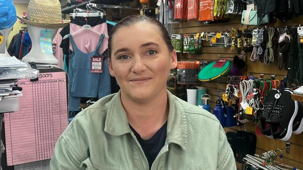 A woman stands in a sports shop. She is wearing a green jacket and a black shirt. Behind her is a collection of different sports apparel. 