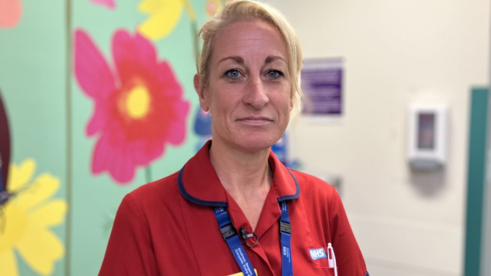 Sally Bryant. She has blonde hair that's tied up and is wearing a red NHS uniform. She is pictured inside a hospital, in front of a wall covered in large flower patterned wallpaper. She is looking at the camera and smiling.