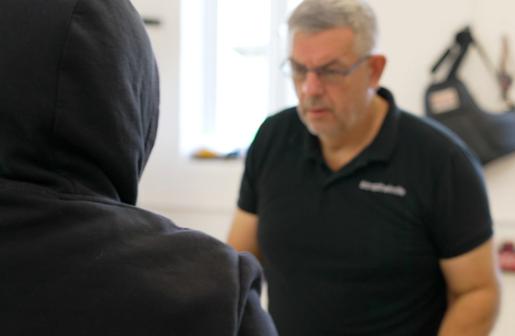 Pete is facing the camera and wearing a black T'shirt and glasses. He is stood in front of Alfie who has a black hooded top on and his hood up with his back to the camera.