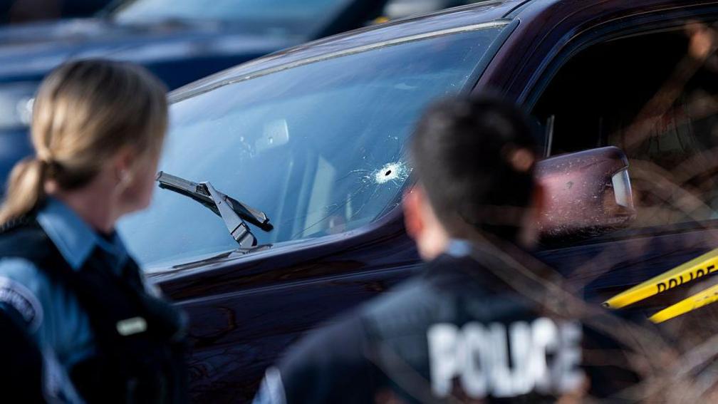 A bullet hole is seen in the windshield of a vehicle involved in a shooting by an ICE agent during federal law enforcement operations on 7 January 2026 in Minneapolis, Minnesota. Two police officers stand in the foreground, out of focus.