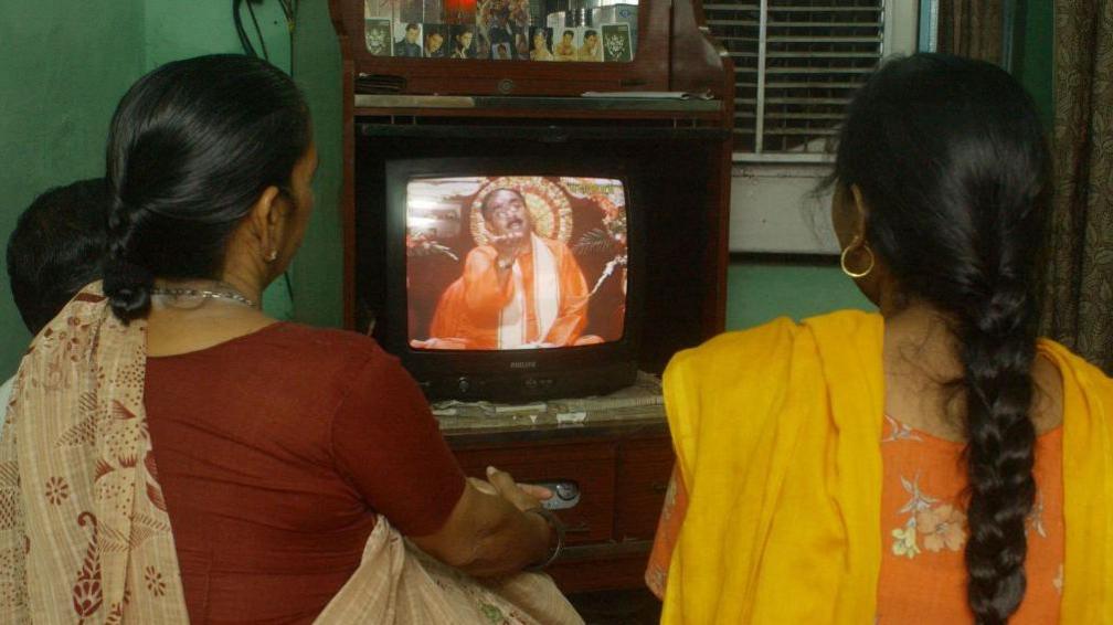 Two unidentified Indian Hindu women watch and listen to a Sadhu (holy man) appearing on a private television channel 'Sadhna', in New Delhi, 03 September 2003. Private television channels like 'Sadna' (Meditation), Aastha (Faith) and Sanskar (Tradition) beam 24-hour Hindu religious programmes to millions of Indian homes, especially in states like Gujarat, Maharashtra and Uttar Pradesh which are undergoing strong Hindu revivalism. (Photo by PRAKASH SINGH / AFP) (Photo by PRAKASH SINGH/AFP via Getty Images)