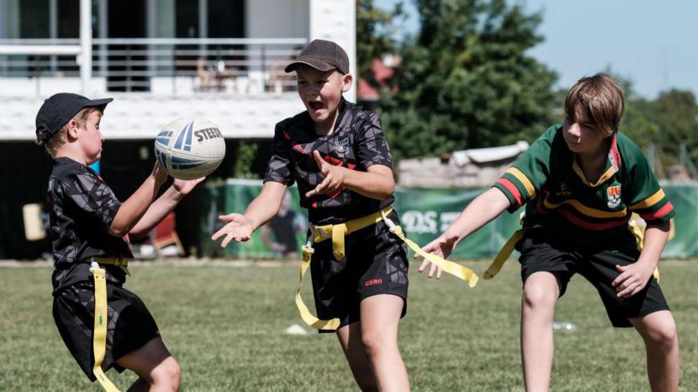 Three children play tag rugby. They have yellow belts with long tags on them and toss a rugby ball between each other. 