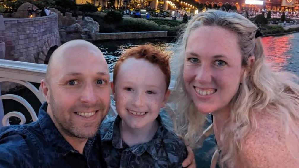 Wayne, Finn and Lisa O'Neil taking a selfie standing on a bridge over a river. There are lights from restaurants and bars in the background.