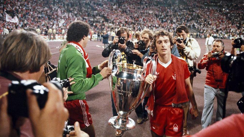 Winning goalscorer Trevor Francis (right) and goalkeeper Peter Shilton parade the trophy after the 1979 European Cup Final between Nottingham Forest and Malmo at the Olympic Stadium on 30 May, 1979 in Munich, Germany. Francis is wearing a red kit and is holding one handle of a giant silver cup. There are crowds in the stadium stands behind the players.