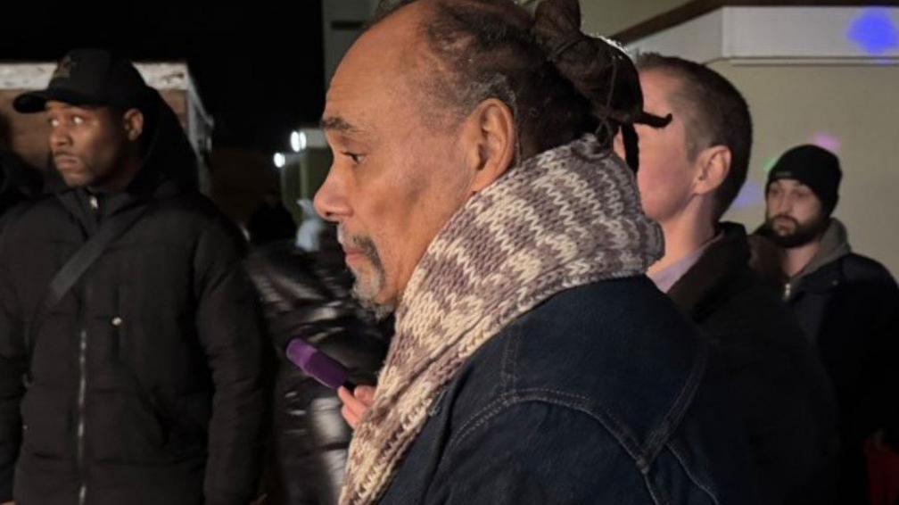 Mr Meade's father, pictured at the vigil, side on to the camera. He is wearing a scarf and other people are in the background