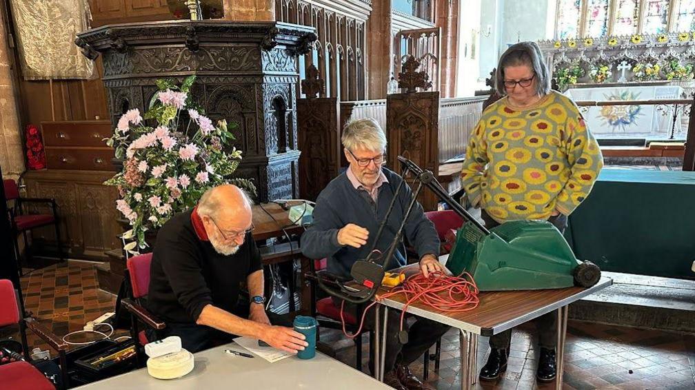Hugh Champion working on a lawn mower, a woman watching what he is doing to fix the lawnmower, and another volunteer working on something else. The two men are sitting at tables.