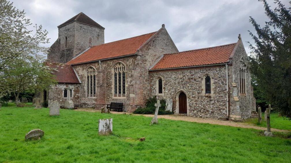 Exterior of All Saints Church in Ashwicken, King’s Lynn, a stone and flint building with ornate arched windows with trees either side and gravestones in front