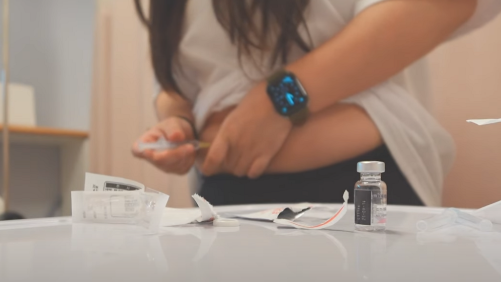 A woman injecting a substance with a syringe on her belly. She is standing in front of a counter, which is strewn with medical products.