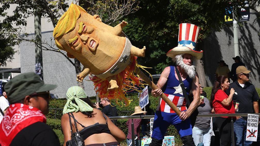 Një burrë i veshur si xhaxhai Sam ka një model të Donald Trump-it kur ishte foshnjë në fund të një shkopi të madh.