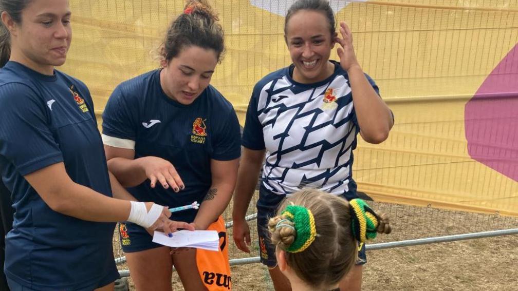 Three female rugby players look happy as they gather around a young player, who is looking at them, to present her with something. They are outside on a field.