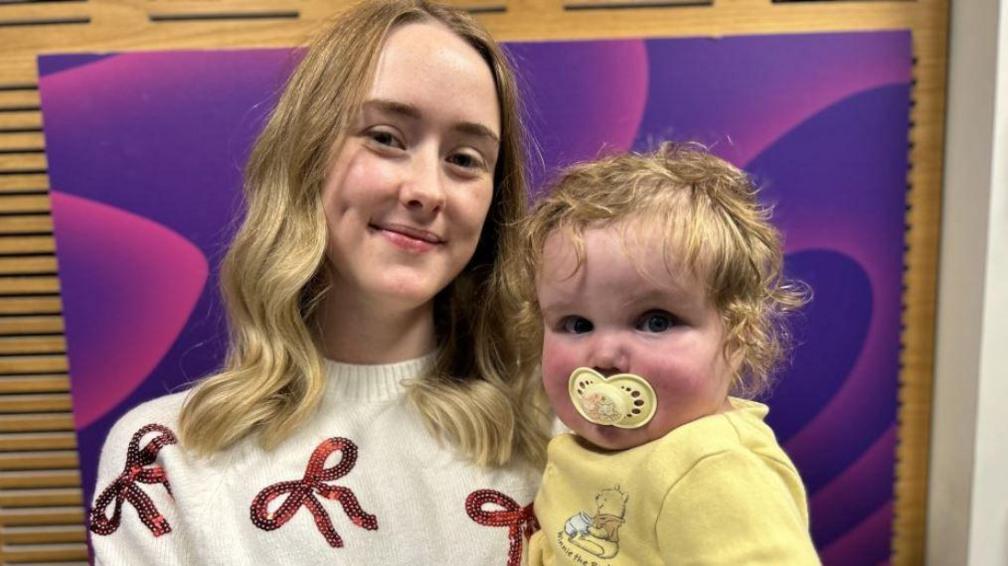 A young mother with long wavy blonde hair in a cream jumper with red sequinned ribbon pattern holding her toddler in front of a swirly purple backdrop. He is blonde and wearing a yellow Winnie the Pooh babygro with a yellow dummy in his mouth.