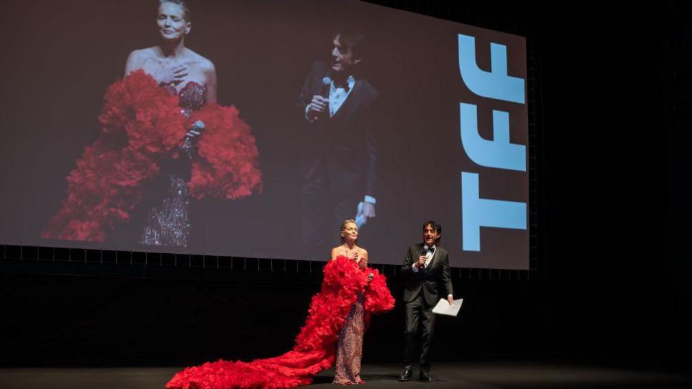 Sharon Stone in a dress with a long red train, speaking in front of a screen showing herself, at the Torino Film Festival, November 2024