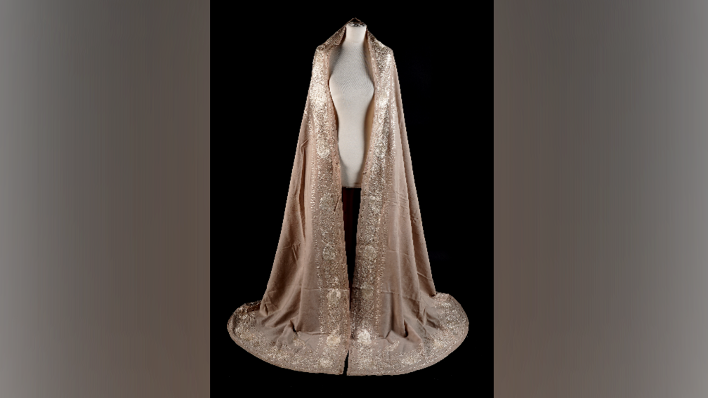 A pink shawl with embroidery of sea shells on the collar and trim, it is on a mannequin in a dark room.