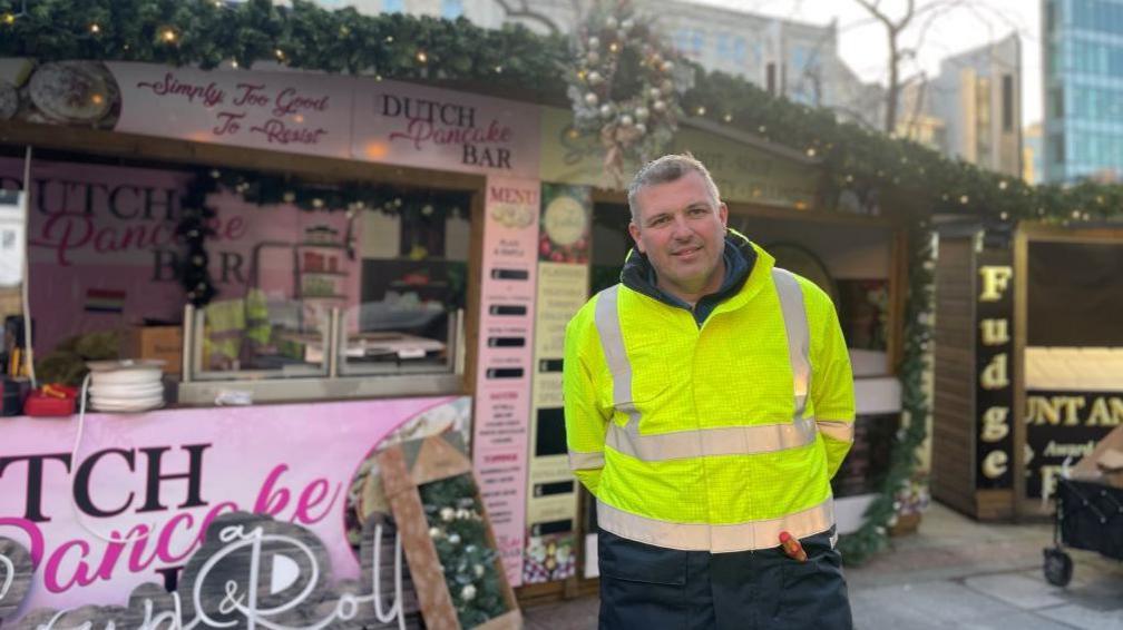 A man in a high-vis coat stands in front of wooden chalet with pink advertising hoarding.